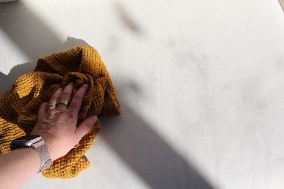 A hand wearing a watch uses a yellow cloth to wipe a white surface with visible smudges and shadows.