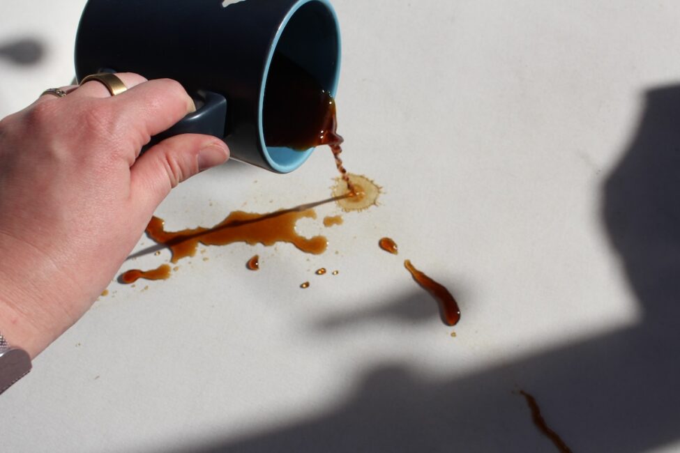 A hand holding a dark blue mug spills coffee onto a white surface, creating several brown stains and drips.