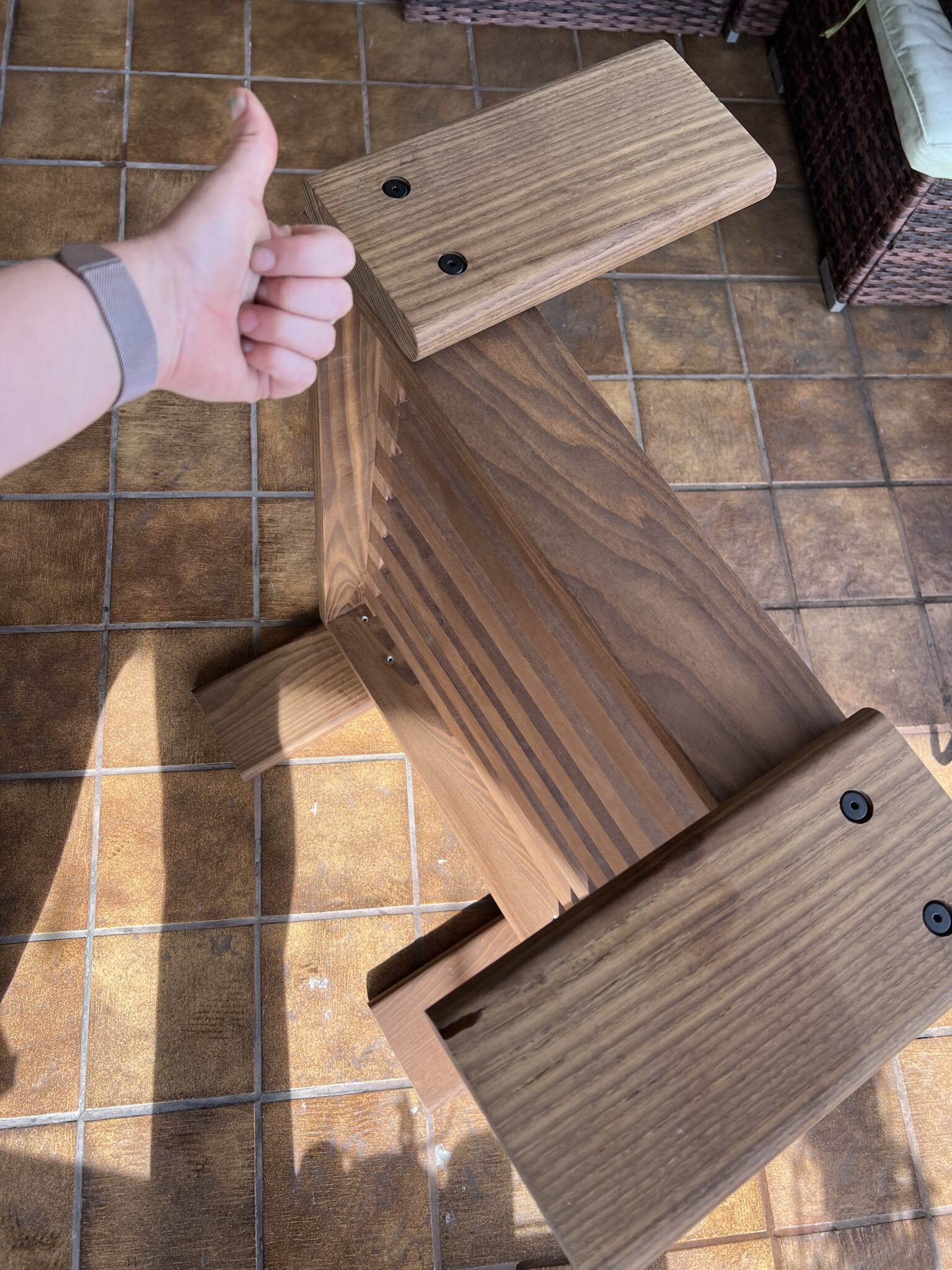 A person gives a thumbs-up next to a wooden chair with a slatted back, placed on a tiled floor in a sunlit room.