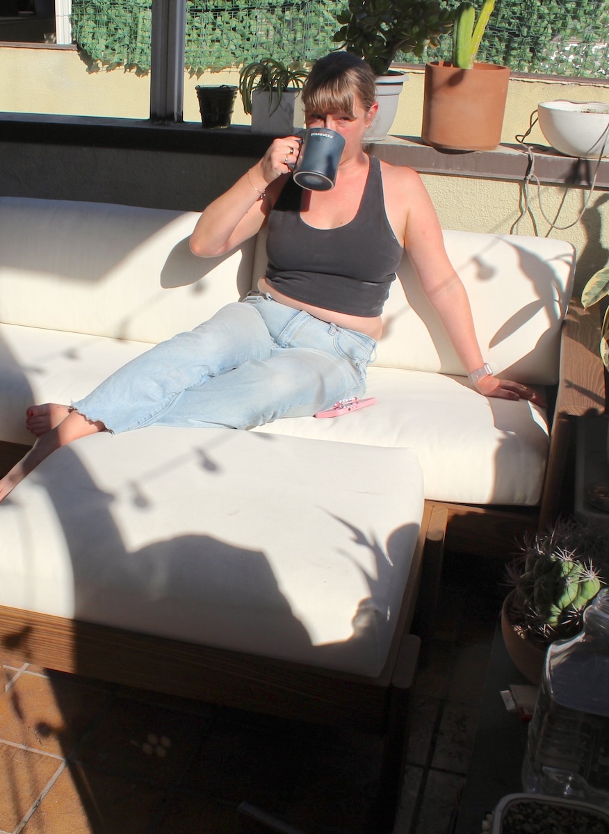 A woman in a black tank top and light jeans sits on a white outdoor sofa, drinking from a mug, with plants and sunlight casting shadows around her.