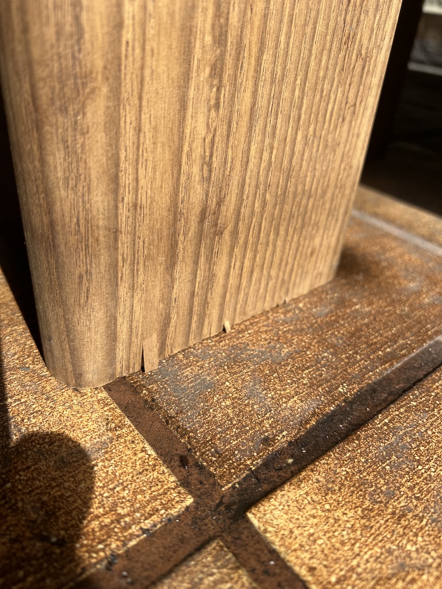 Close-up of a wooden table leg with two visible cracks or splits at its base, standing on a textured wooden floor in bright sunlight.