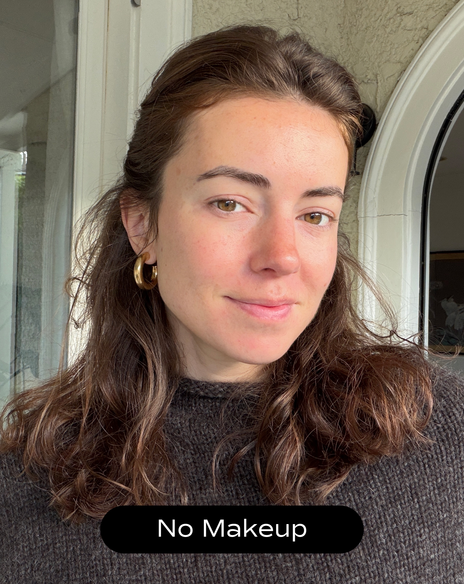 A woman with wavy brown hair and gold hoop earrings, wearing a dark sweater, stands indoors with a neutral expression. Text over image reads "No Makeup.