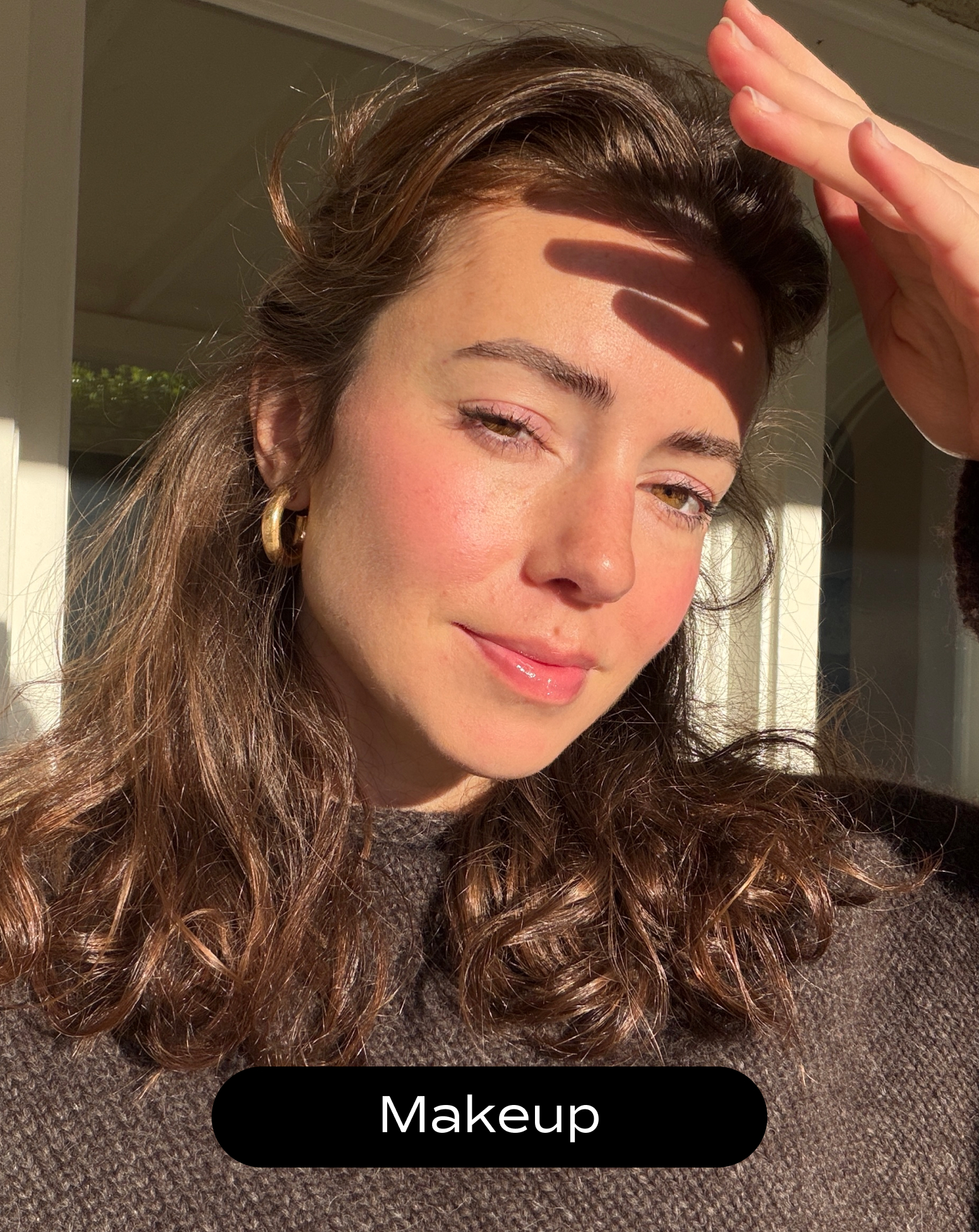 A woman with wavy brown hair and gold hoop earrings poses in sunlight, casting a shadow on her face. She wears natural makeup and a brown sweater. The word "Makeup" appears at the bottom.