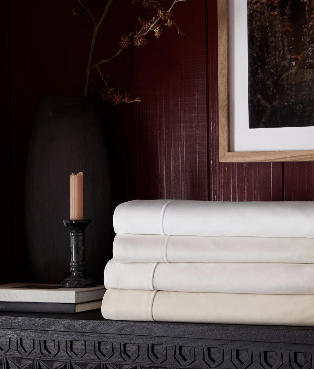 Three neatly folded sheets in light neutral colors sit on a dark wooden surface beside a candle, books, a vase with branches, and a framed picture on a red wall.
