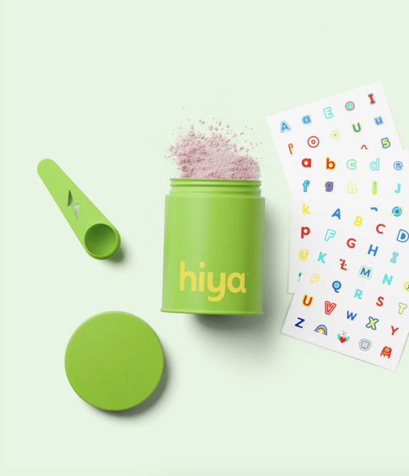 A green container labeled "hiya" with pink powder inside, a matching scoop, a detached lid, and a colorful alphabet sticker sheet on a light green background.