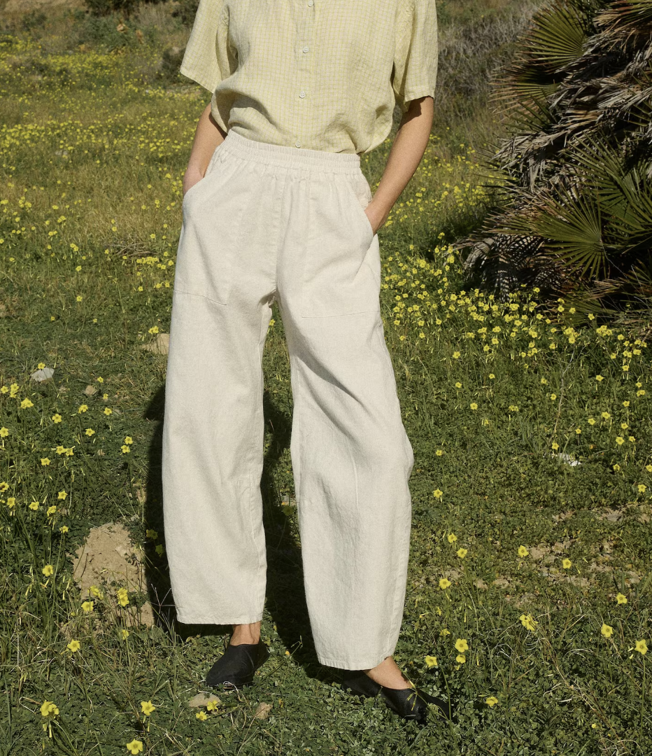 Person standing outdoors in a field of yellow flowers, wearing a light yellow checkered shirt, loose white pants, and black shoes.
