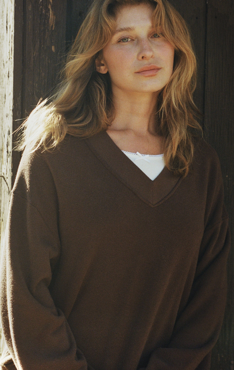 A woman with wavy, light brown hair stands in front of a wooden wall, wearing a brown V-neck sweater over a white shirt and looking calmly at the camera.