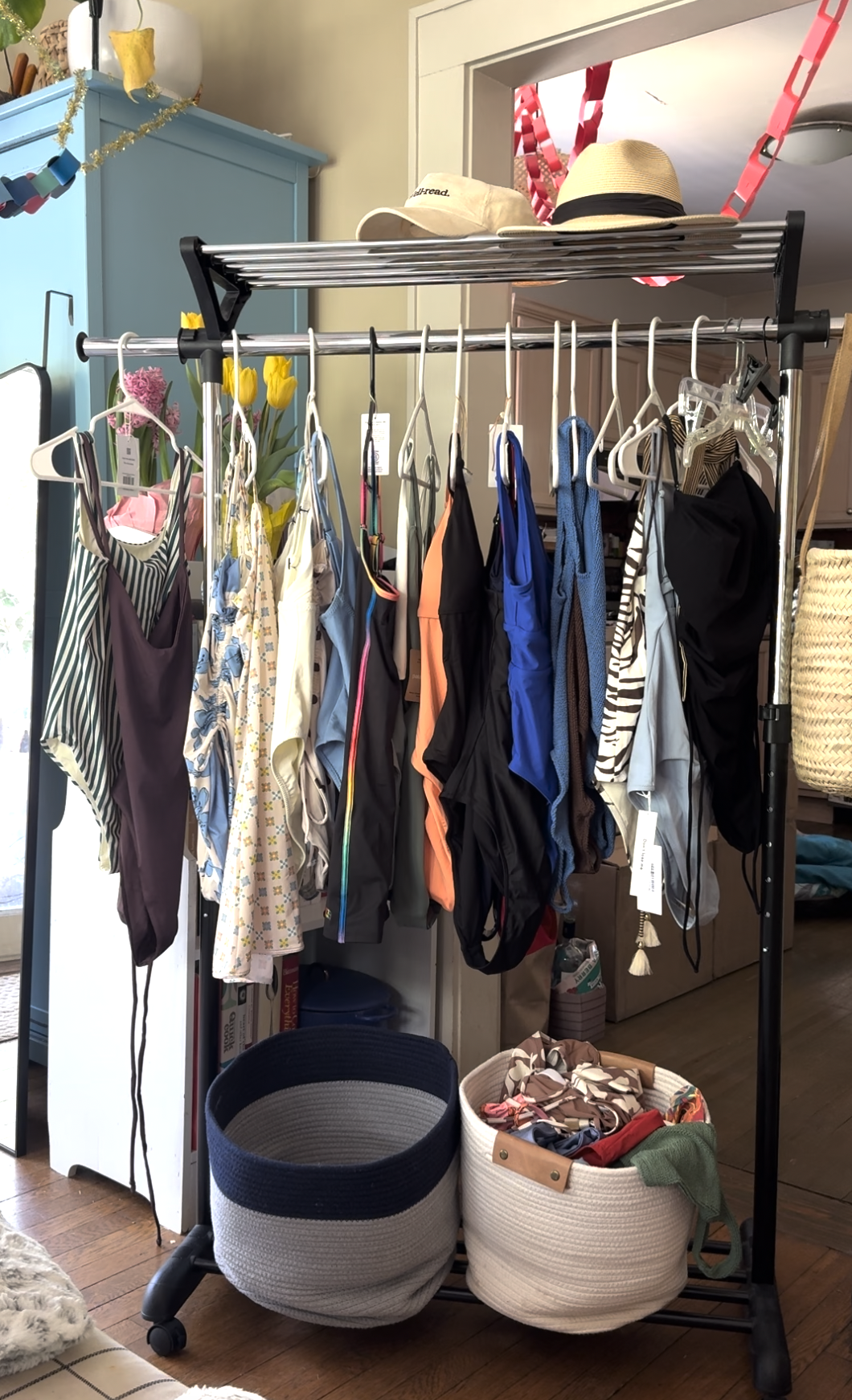 A clothing rack displays various swimsuits and hats, with two baskets full of accessories and items on the floor beneath it in a sunlit room.