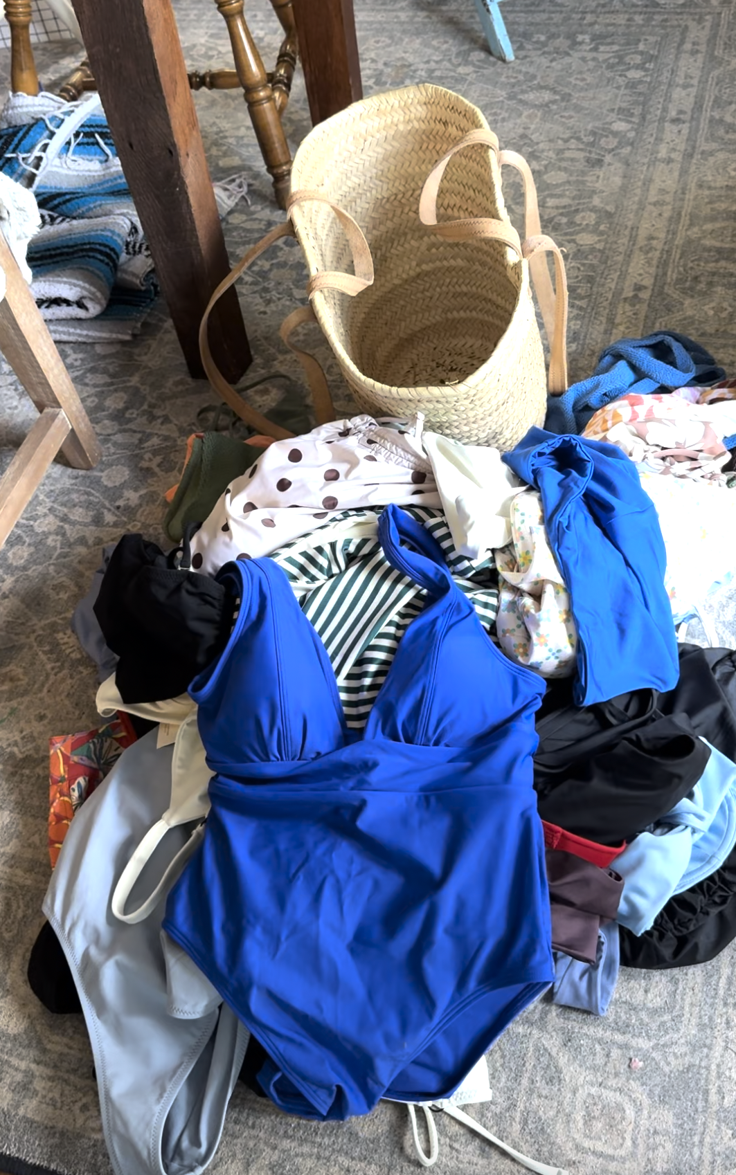 A straw tote bag sits on the floor next to a pile of assorted swimsuits and clothing, with various colors and patterns visible.