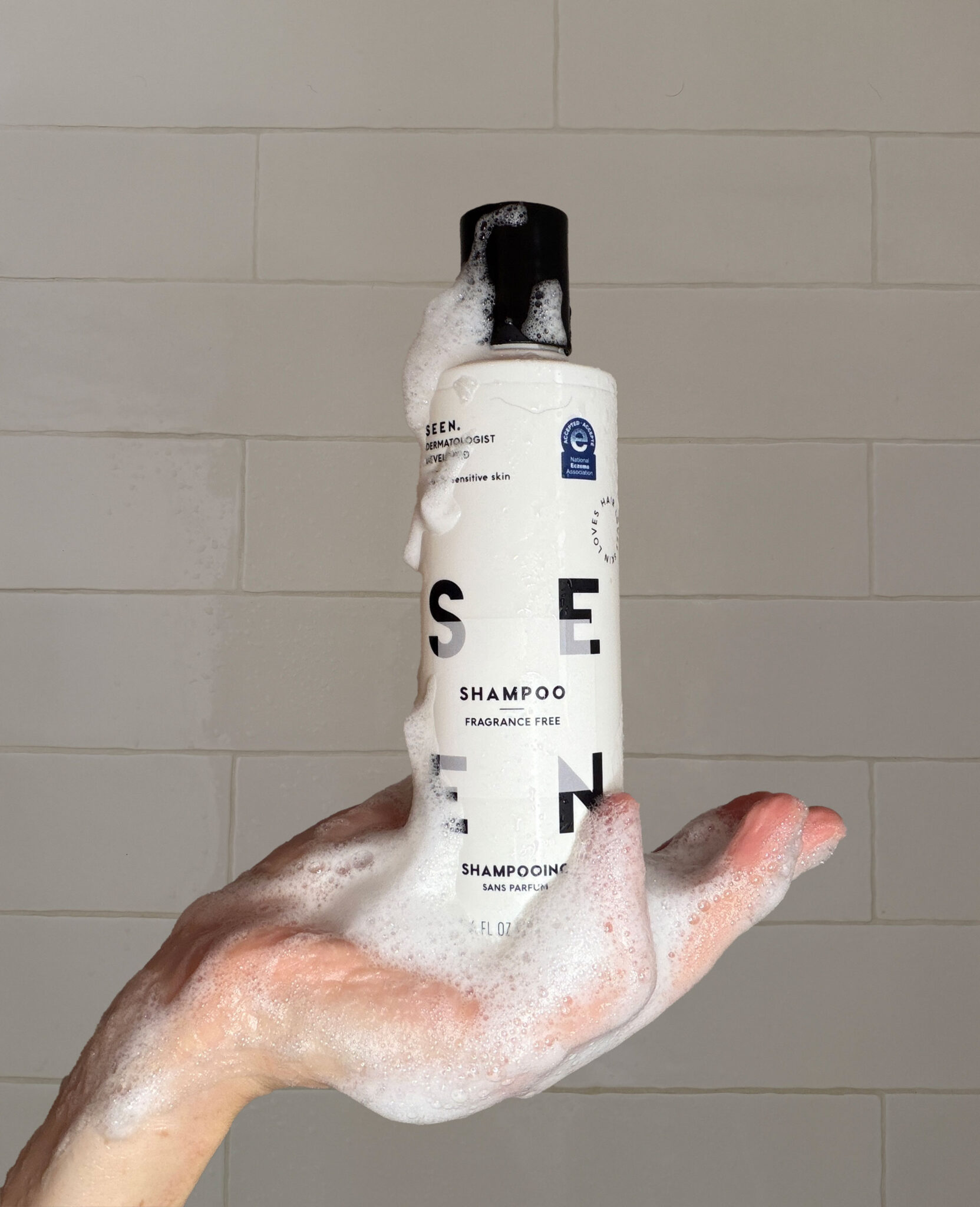 A hand covered in soap suds holds a white bottle of fragrance-free shampoo against a tiled shower wall.