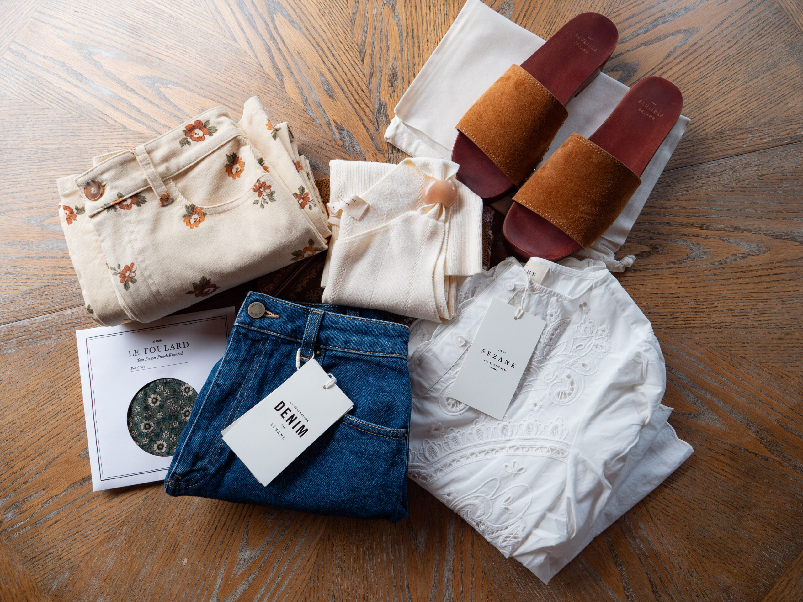 Folded clothes, a pair of brown sandals, a scarf in packaging, and a cream tote bag are arranged on a wooden surface.