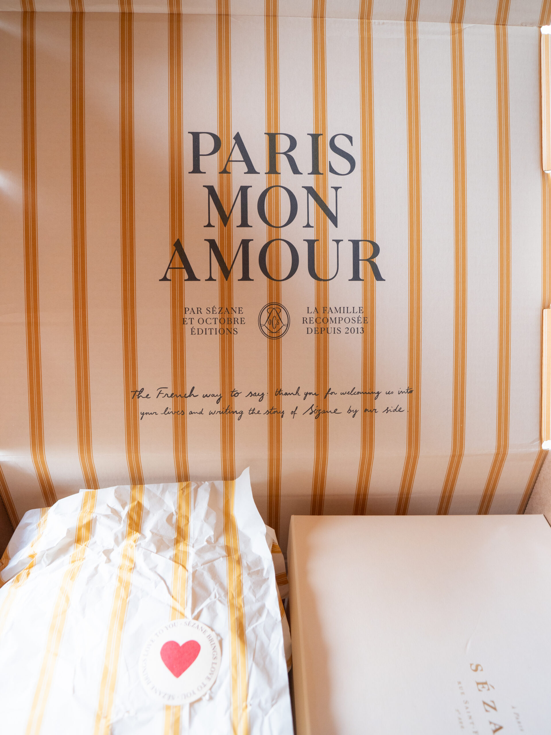 A box with vertical yellow stripes, containing a wrapped item, an envelope, and the words "PARIS MON AMOUR" and a short printed message on the inner lid.
