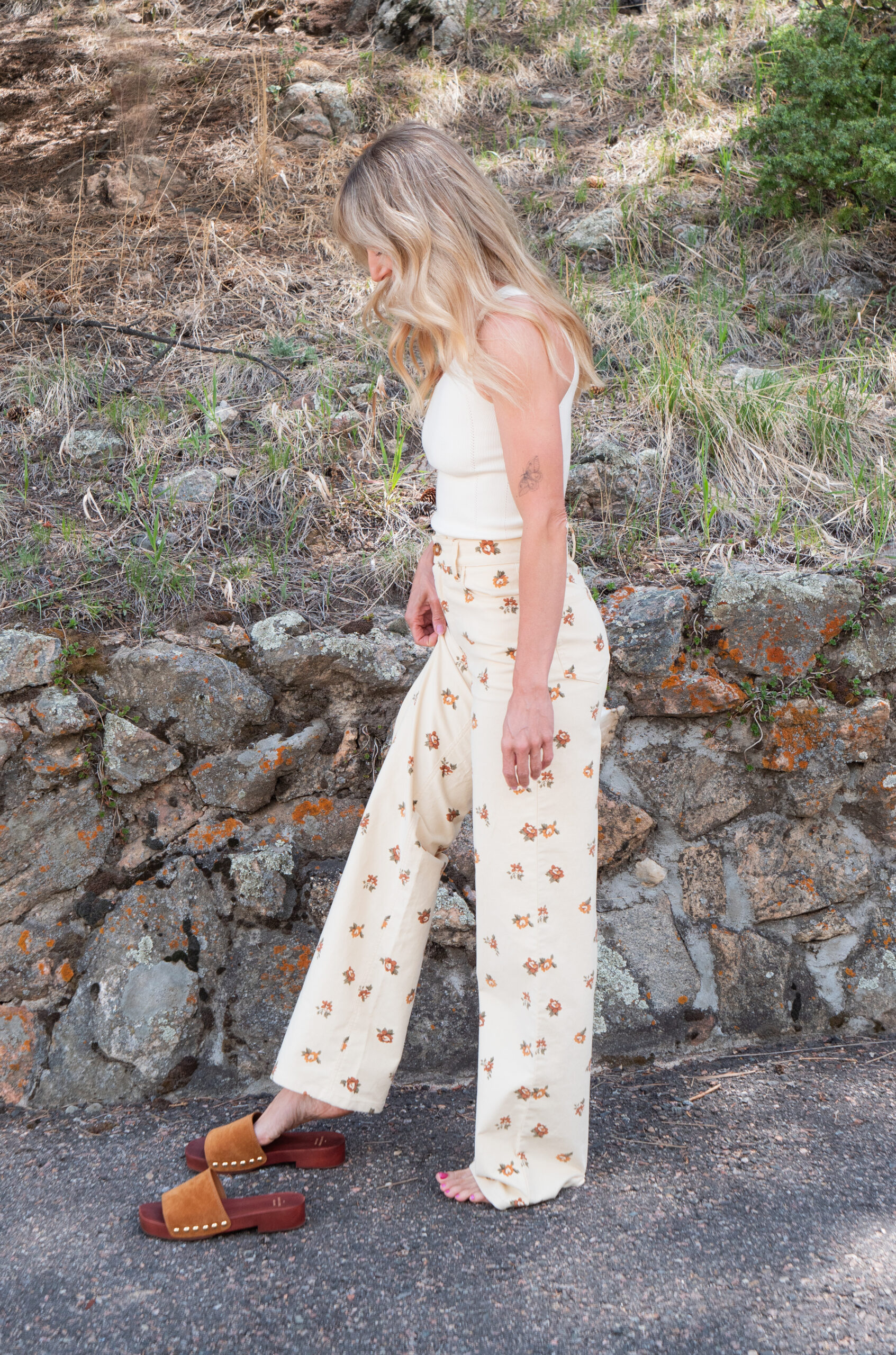 A woman with blonde hair, wearing a white sleeveless top and cream-colored floral pants, stands barefoot on pavement next to brown sandals, with a stone wall and greenery in the background.