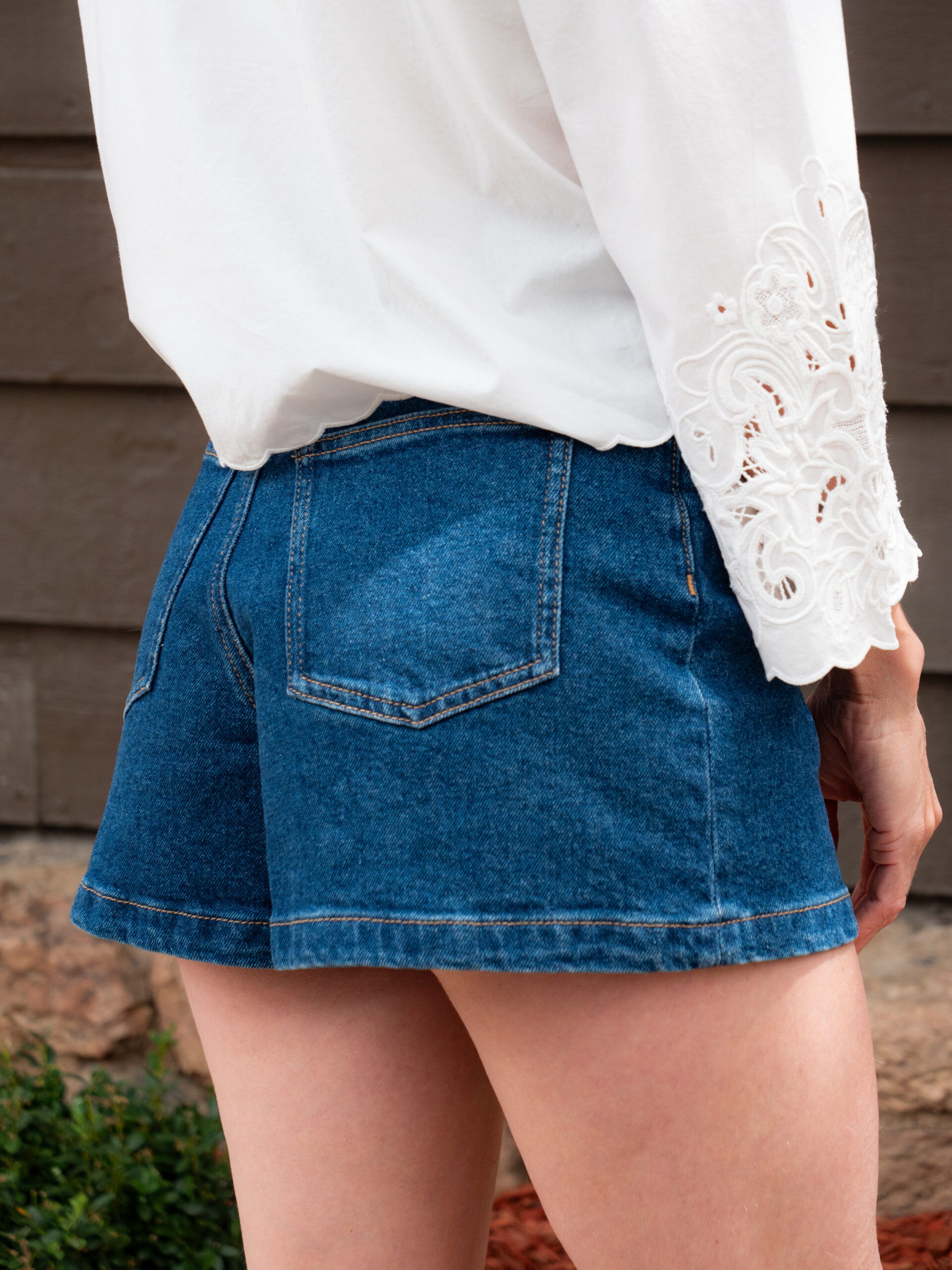 A person wearing a white long-sleeve blouse with lace details and blue denim shorts stands outdoors near a brown wall.