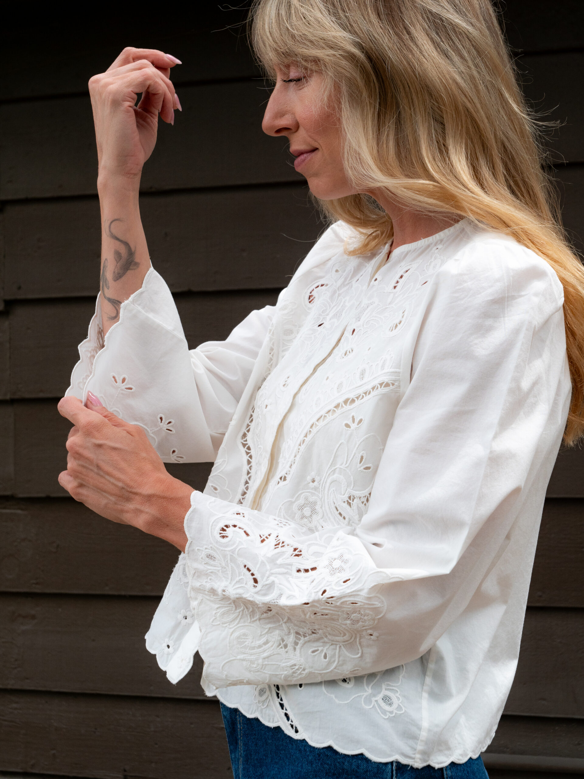 A person with long blonde hair adjusts the cuff of a white, embroidered blouse while standing in front of a dark wooden wall.
