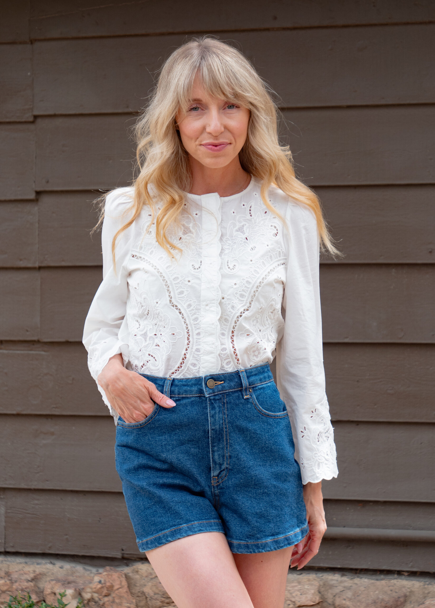 Woman stands in front of a brown wooden wall, wearing a white long-sleeve blouse with lace details and blue denim shorts.