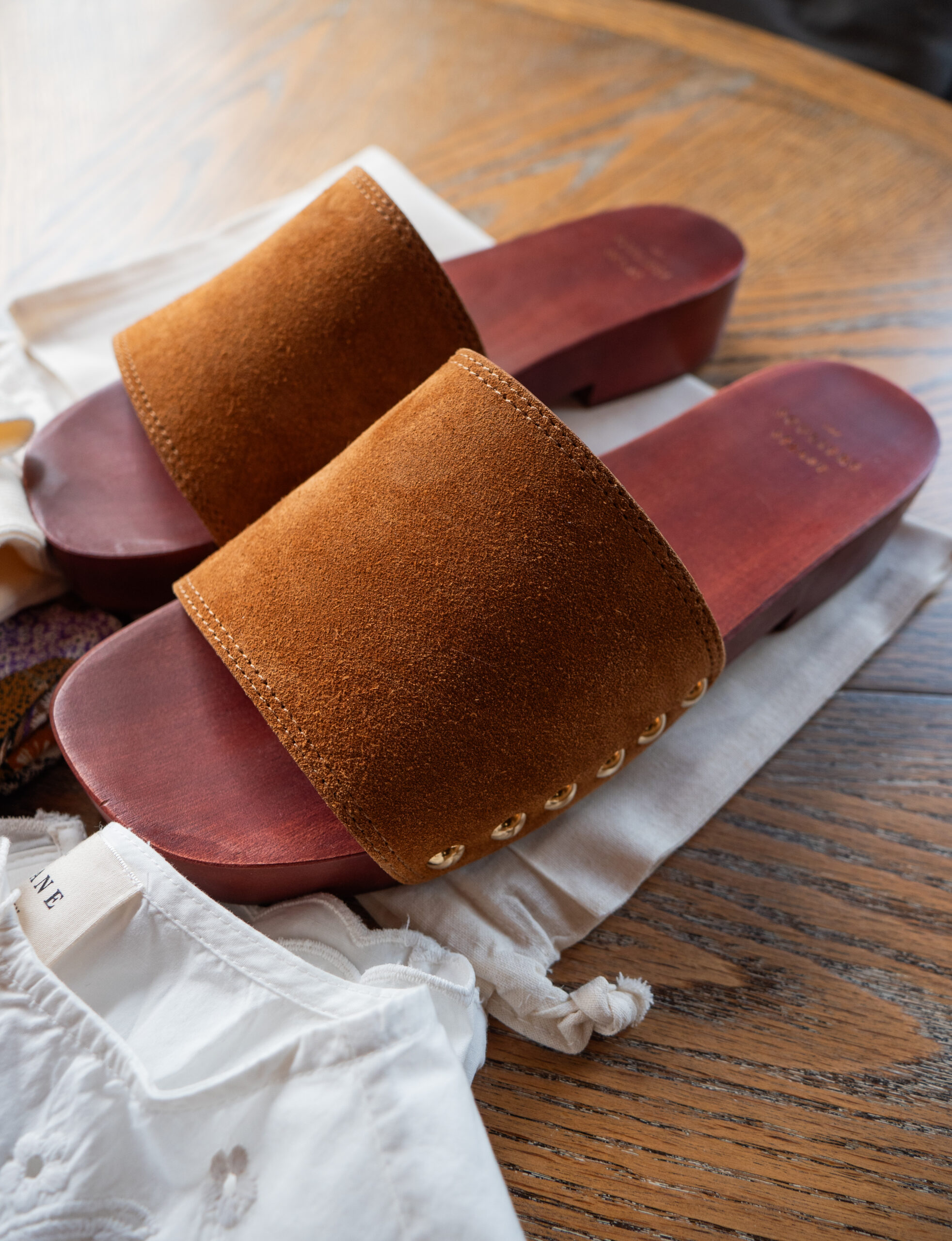 A pair of brown suede slide sandals with wooden soles, resting on a white cloth bag atop a wooden table, next to a white garment.