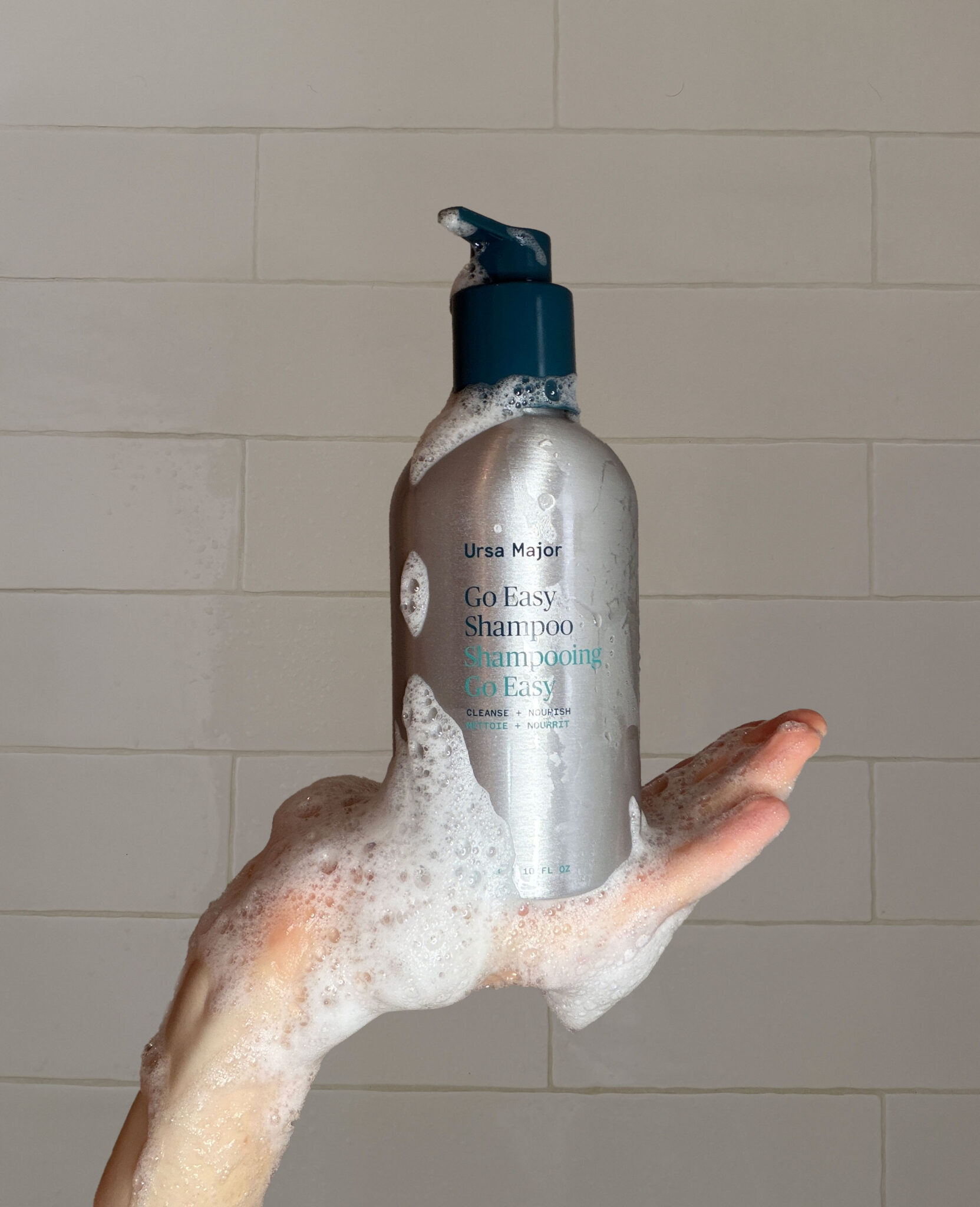 A hand covered in soap suds holds a bottle of Ursa Major Go Easy Shampoo against a tiled shower wall.