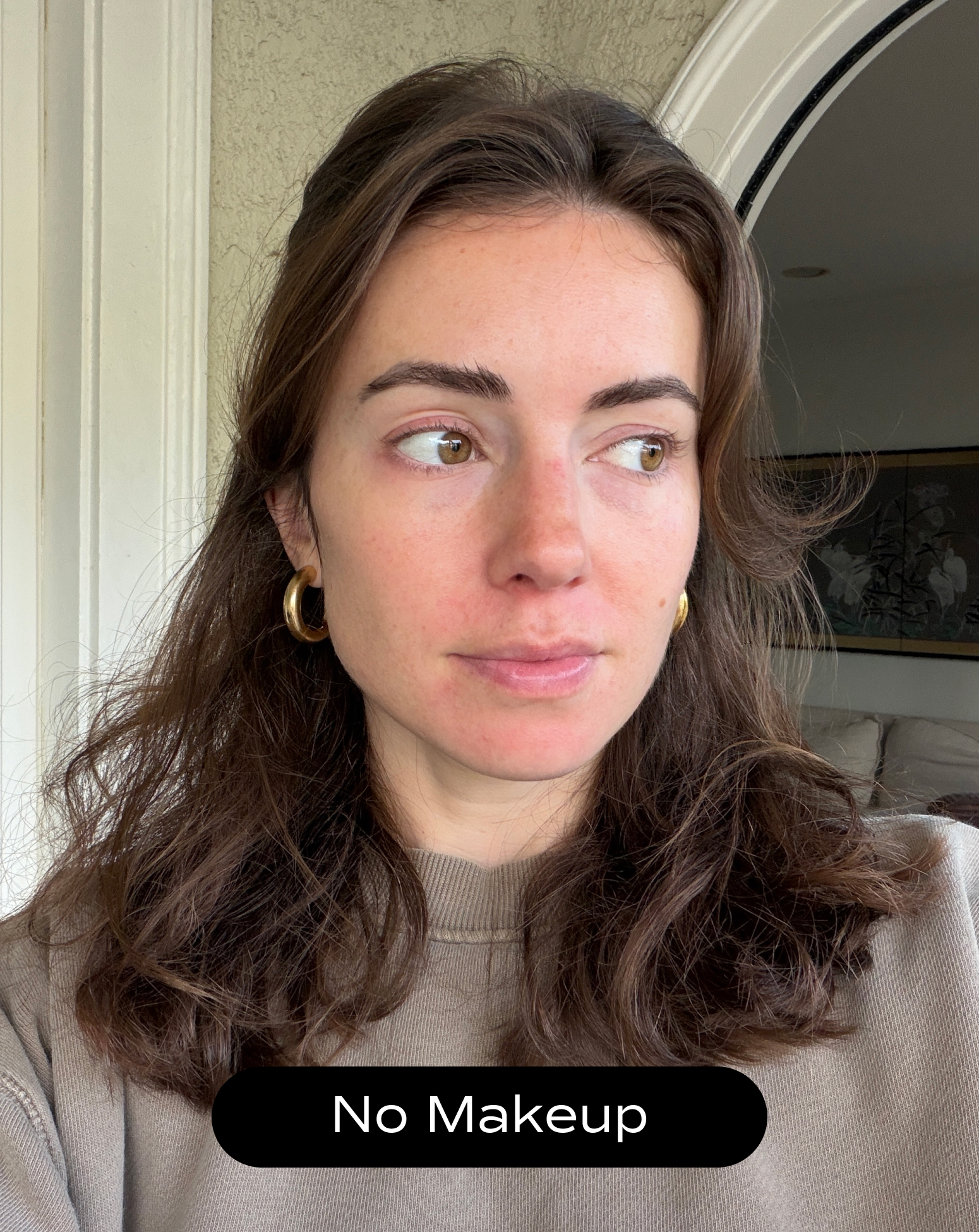 A woman with wavy brown hair and gold hoop earrings looks to the side indoors. She is wearing a beige sweater and has no makeup on. Text at the bottom reads, "No Makeup.