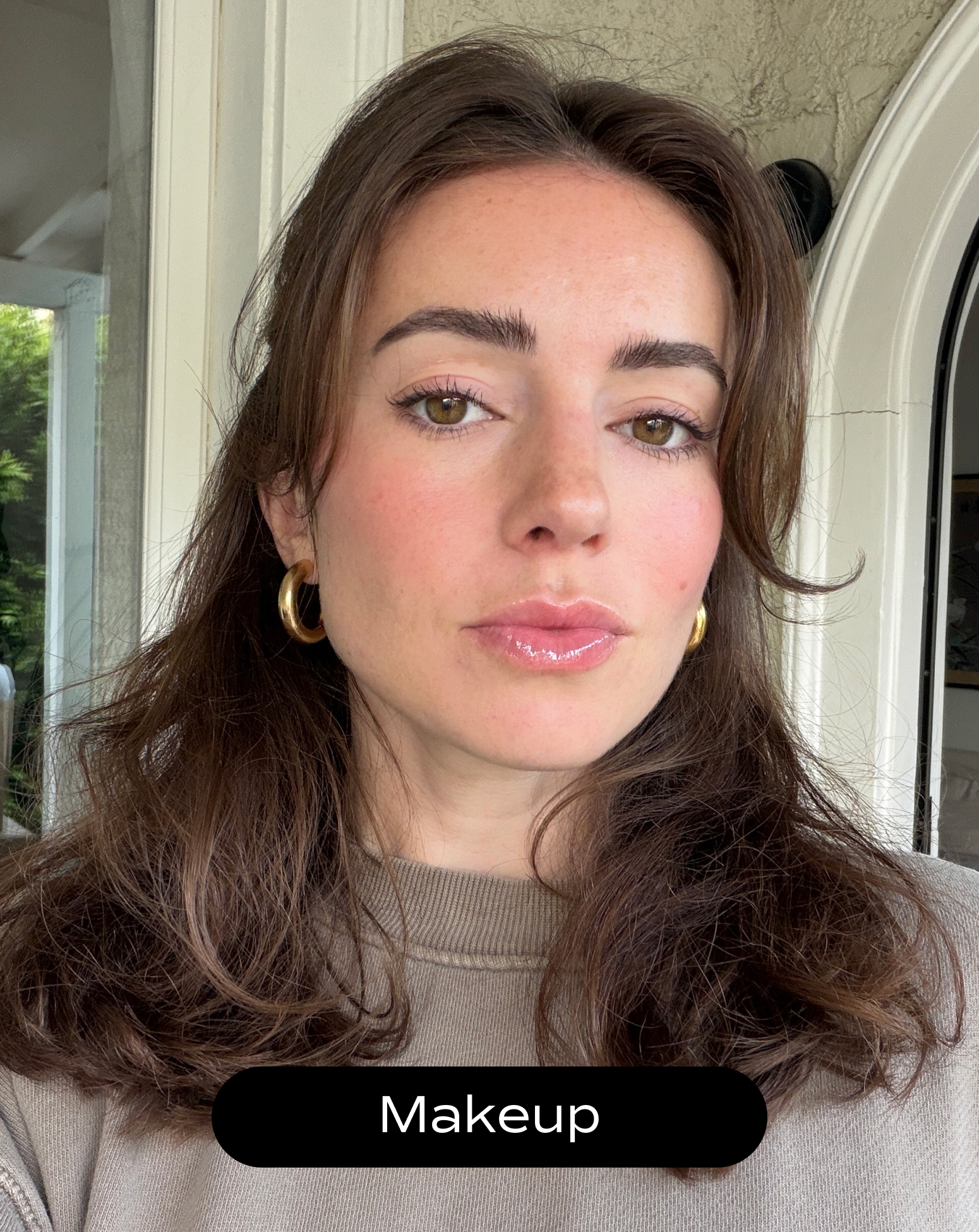 A woman with wavy brown hair, gold hoop earrings, and light makeup including lip gloss and blush poses indoors near a window. The word "Makeup" appears at the bottom.