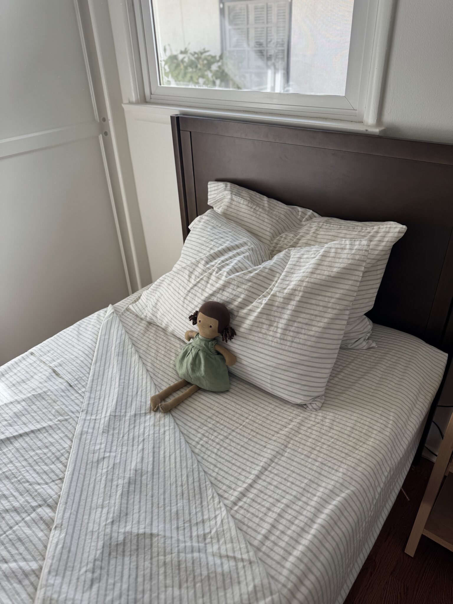 A neatly made single bed with striped white bedding and a fabric doll in a green dress placed on the pillow, beside a window with natural light.