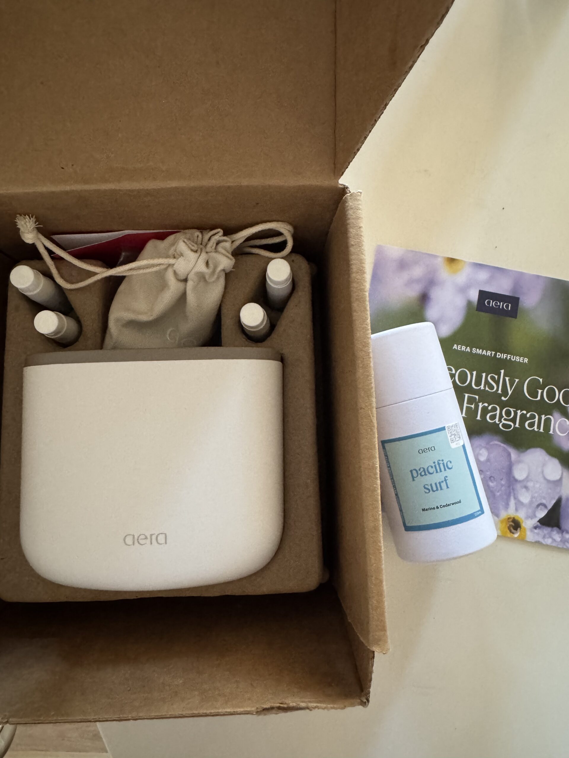 Open box with an Aera smart diffuser, four small fragrance vials, a drawstring pouch, and a “Pacific Surf” scent bottle next to an Aera product card.