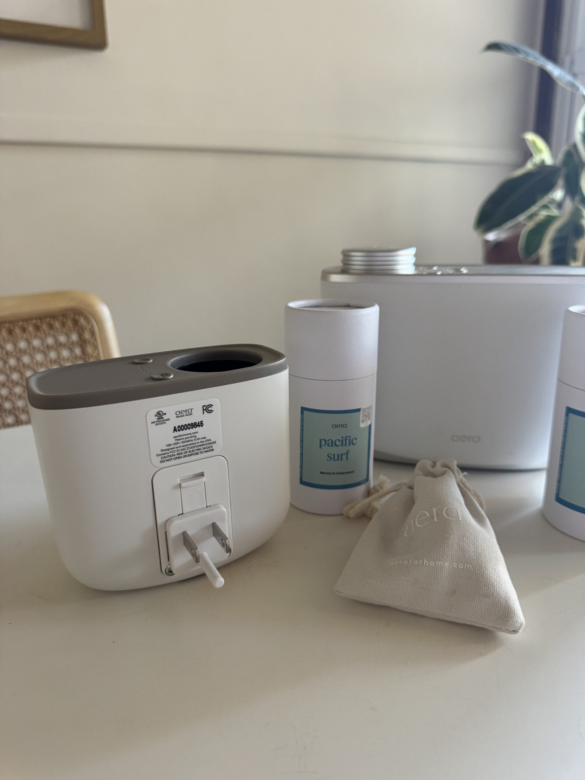 A white countertop holds a plug-in diffuser, two cartridge containers labeled “Pacific Surf,” a cloth pouch, and a plant in the background.