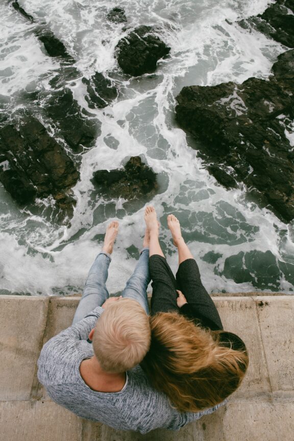 Two people sit side by side on a concrete ledge above rocky ocean waves, with their feet dangling over the edge.