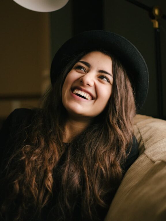 A woman with long brown hair and a black hat sits on a beige couch, smiling and looking upward. The background is softly lit indoors.