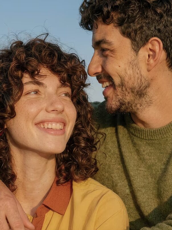 A woman and a man sit close together on a bench outdoors, smiling and looking at each other, with the sky in the background.
