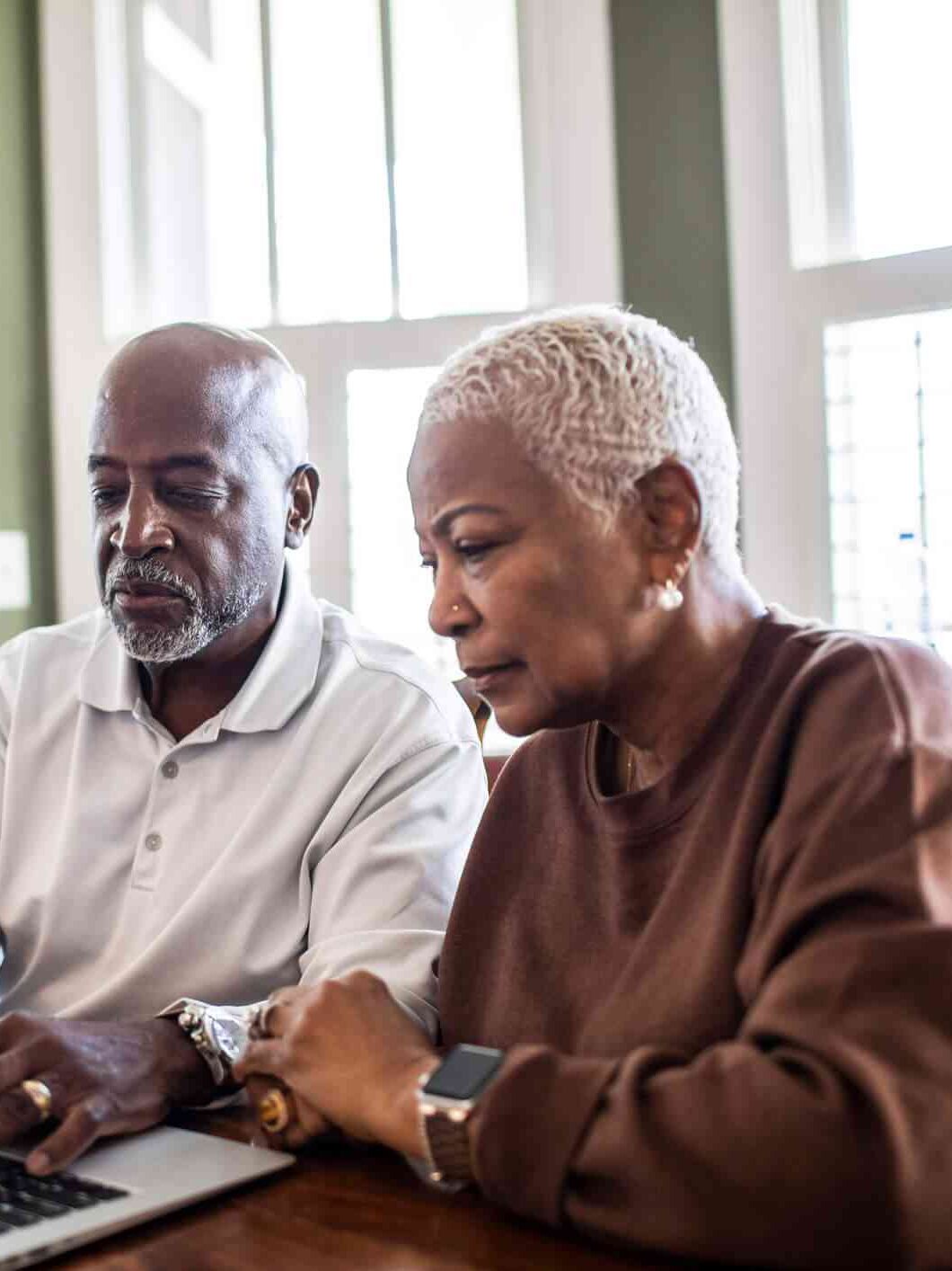 Two older adults sit at a wooden table, looking intently at a laptop computer in a well-lit room with large windows.
