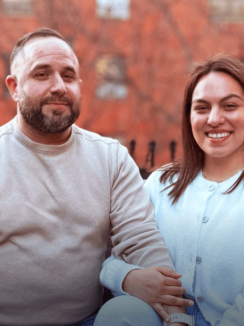 A man and woman sit closely together outdoors, both smiling at the camera. The background shows a blurred brick wall and bare tree branches.
