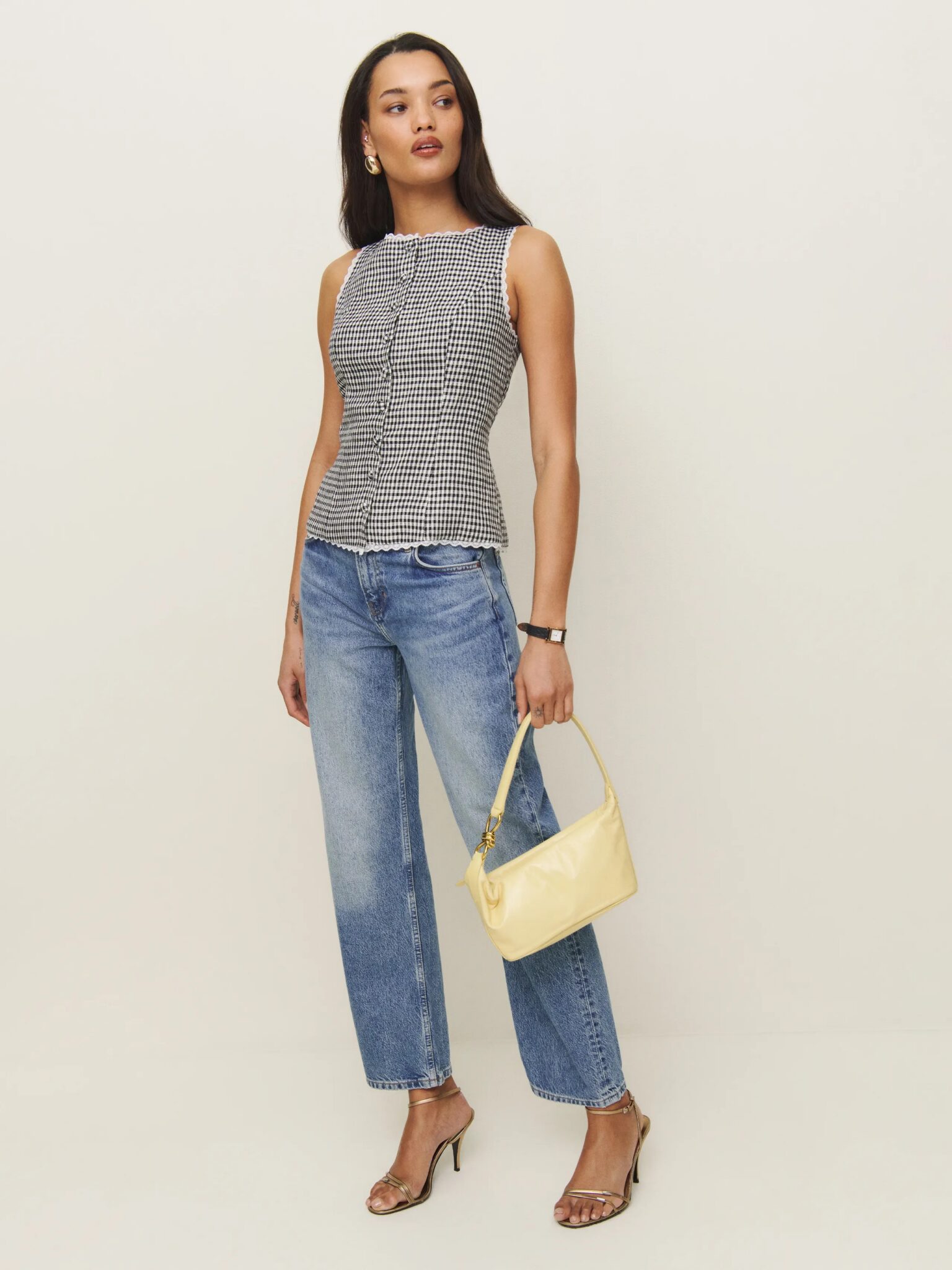 A woman stands against a plain background wearing a sleeveless checkered top, blue jeans, heeled sandals, and holding a small yellow handbag.