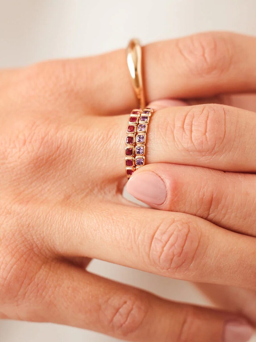 Close-up of a hand wearing two gold rings, one simple band and one with a double row of small red gemstones, against a neutral background.