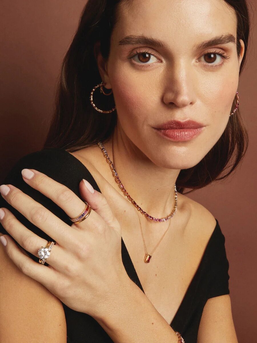 A woman with long brown hair wears a black top, gold hoop earrings, a gold necklace, and multiple rings, posing with one hand resting on her shoulder against a brown background.