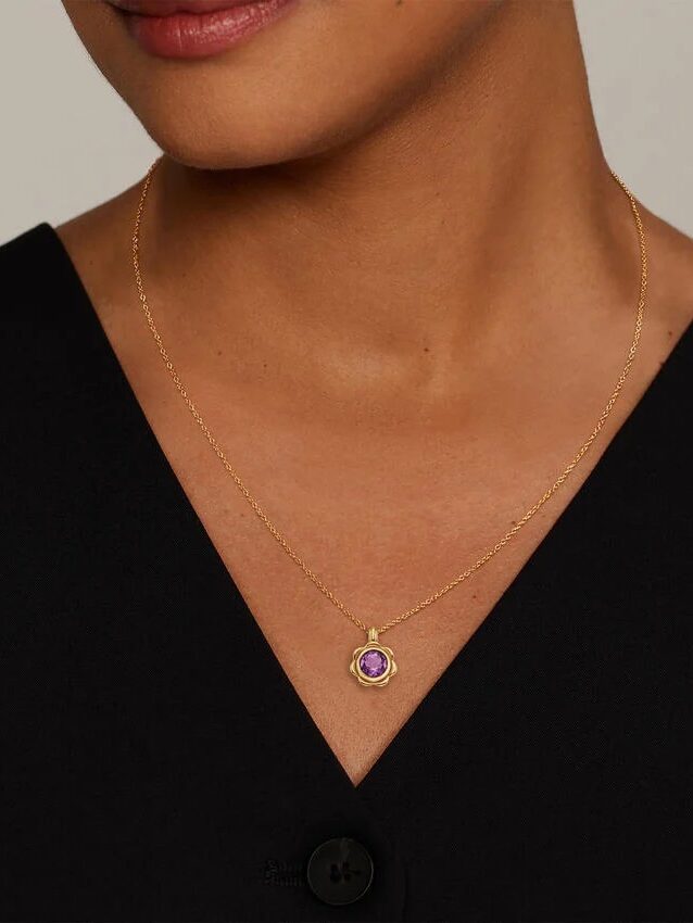 A woman wearing a black V-neck top and a gold necklace with a round purple gemstone pendant.