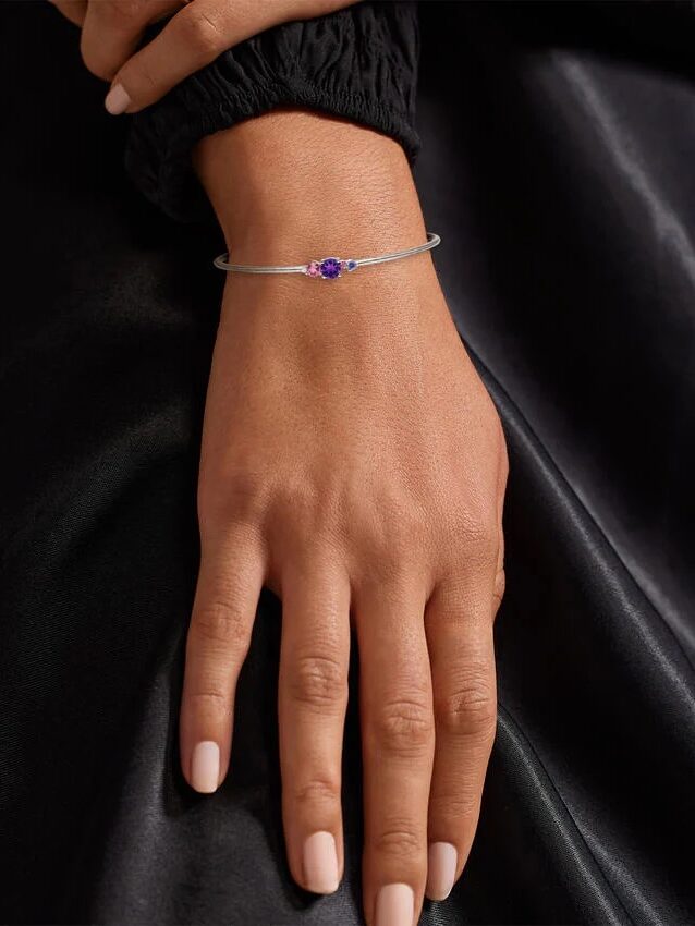 A person rests their hand on a dark satin surface, wearing a thin silver bracelet with a small purple gemstone.