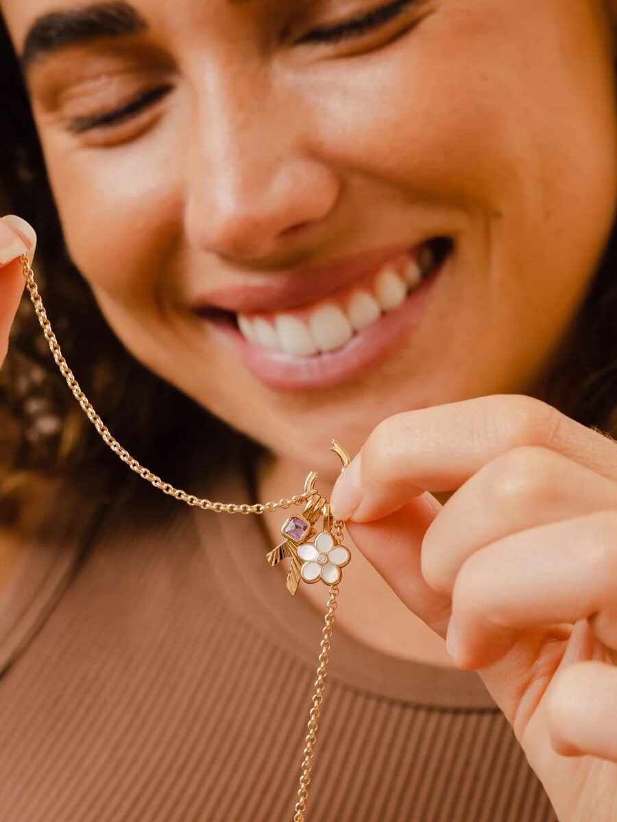 A woman smiles while holding a gold necklace with a pendant featuring a white flower and a small purple stone.