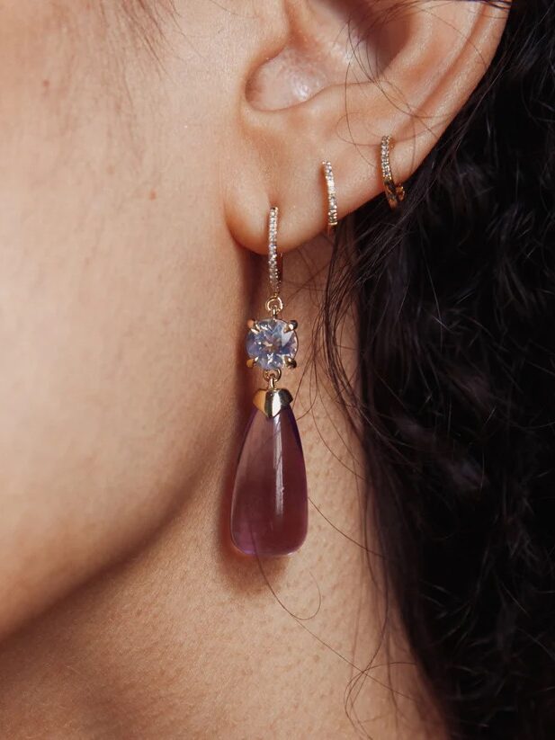 Close-up of a person's ear wearing three small gold hoop earrings and a larger dangling earring with a purple teardrop-shaped pendant and a clear gemstone.