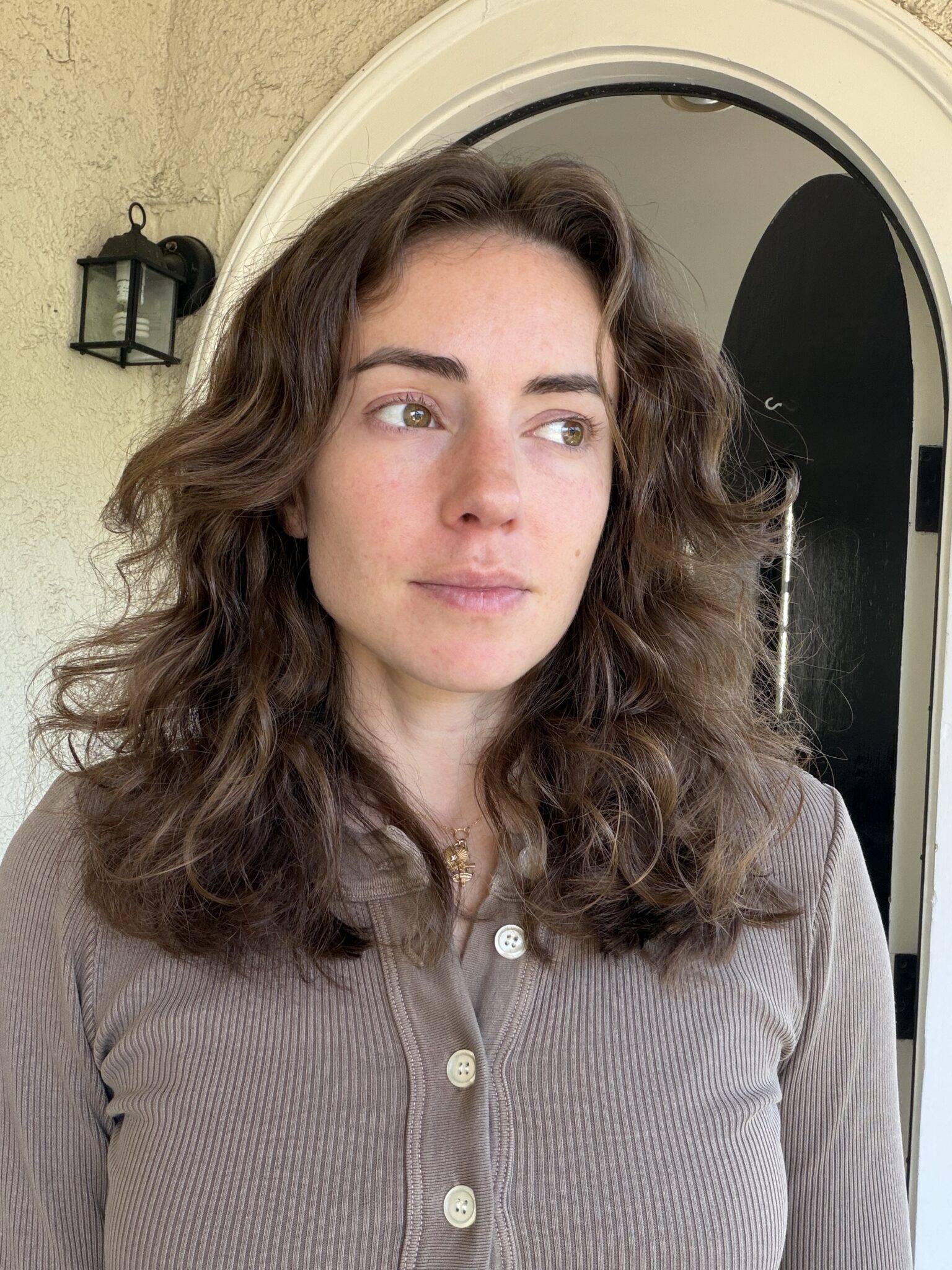A woman with wavy brown hair and a neutral expression stands in front of an arched doorway, wearing a taupe button-up top.