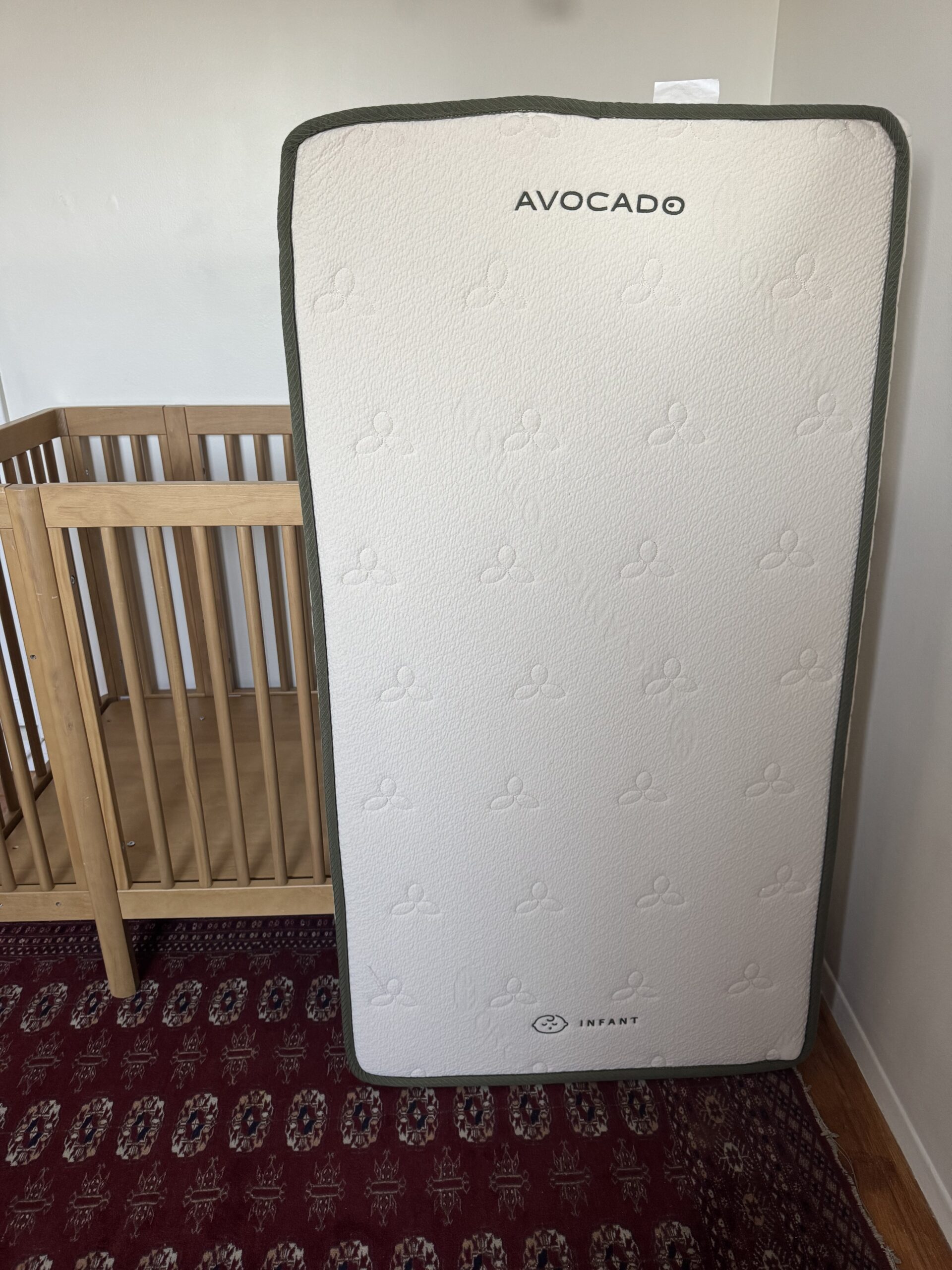 A crib mattress labeled "Avocado" stands upright next to a wooden crib on a red patterned rug in a room with white walls.