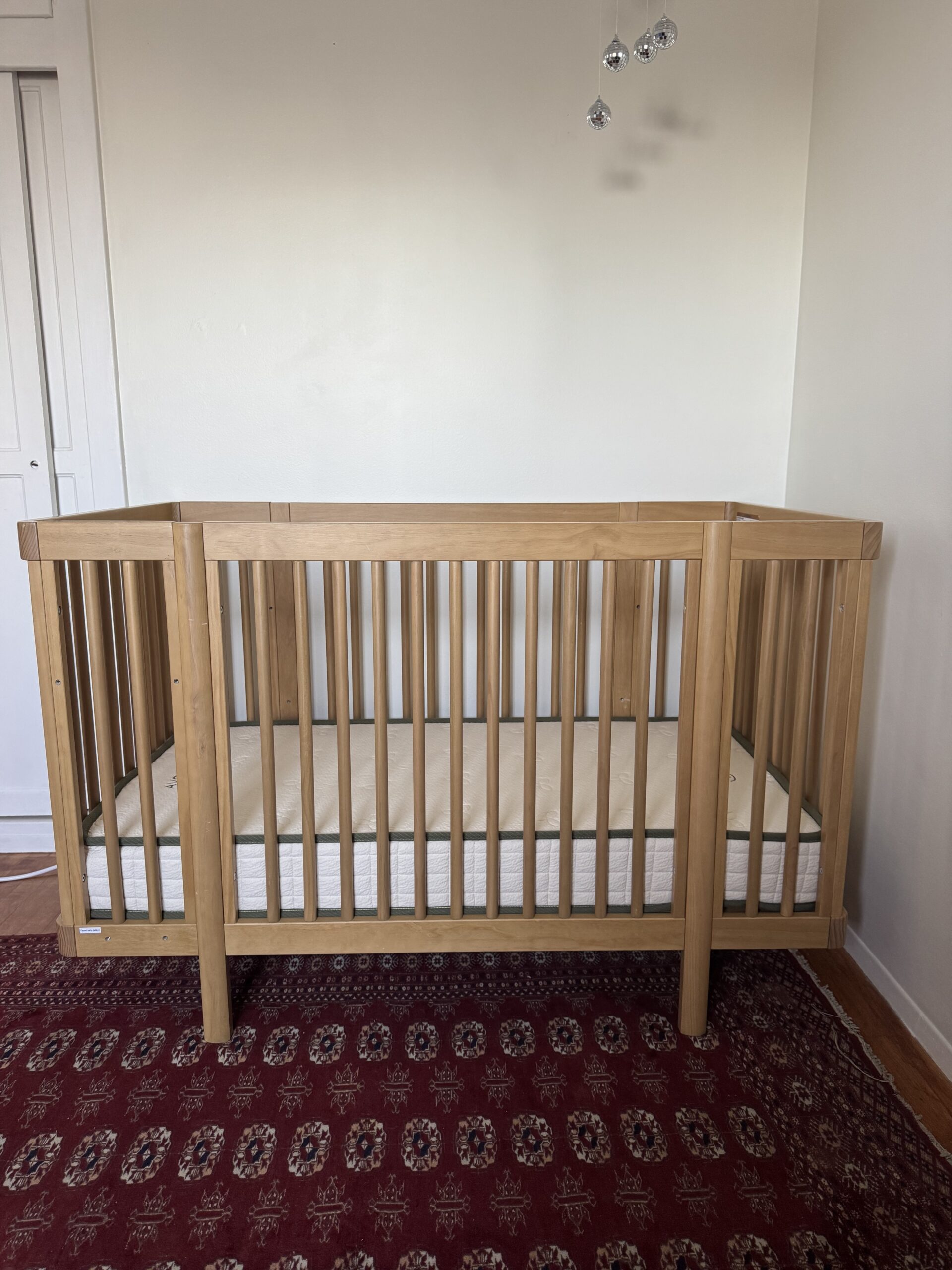 A wooden baby crib with a mattress stands on a patterned red rug against a plain white wall.