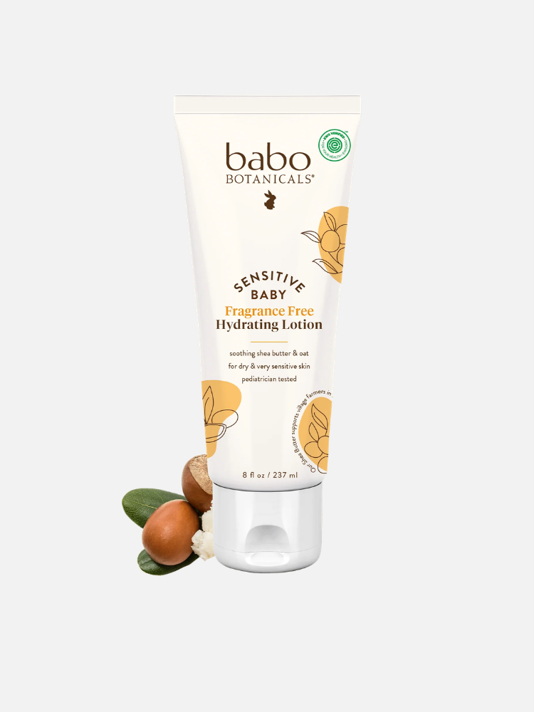 A tube of Babo Botanicals Sensitive Baby Fragrance Free Hydrating Lotion, 8 oz, shown with shea nuts and leaves beside it.