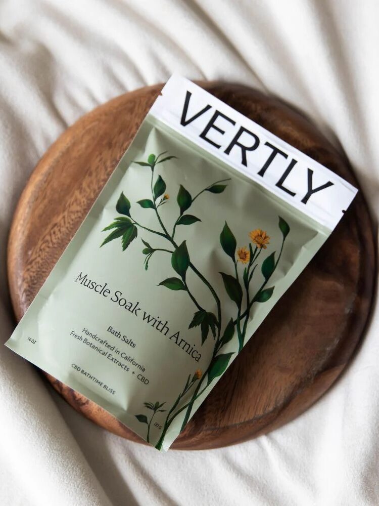 A Vertly Muscle Soak with Arnica packet rests on a round wooden tray placed on a white fabric surface.