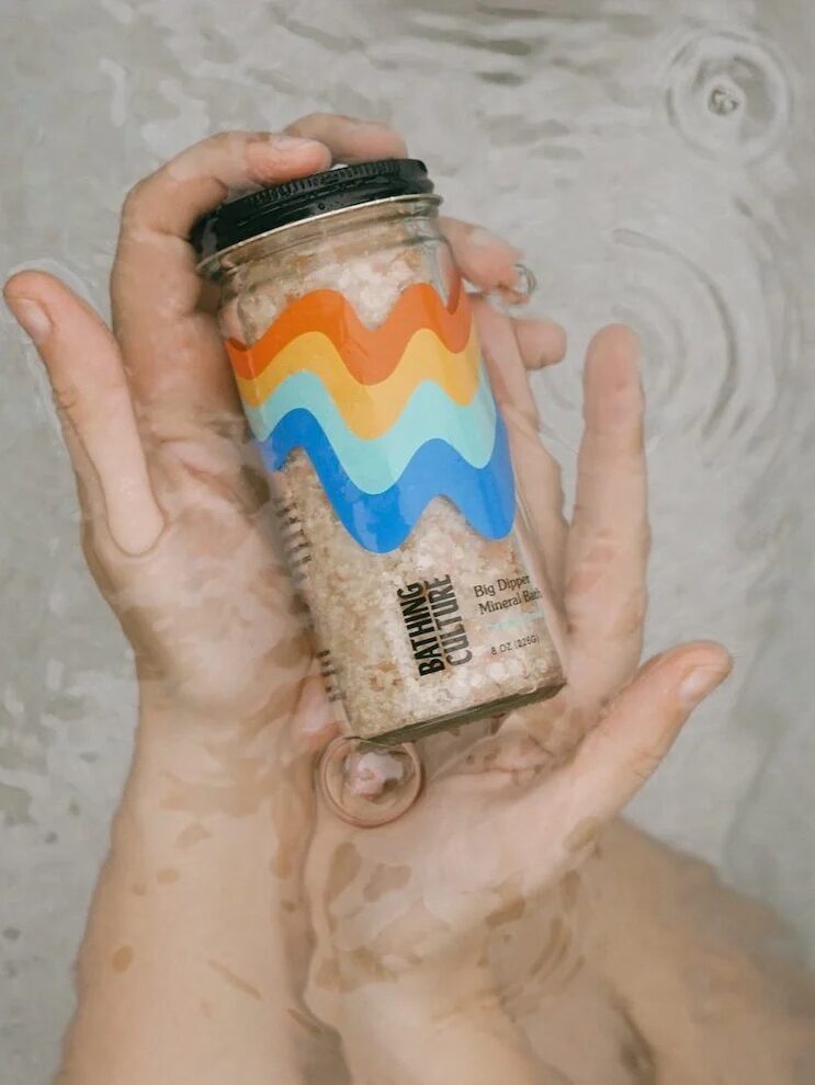 A person holds a clear jar with a colorful wavy label underwater; ripples and reflections are visible on the surface.