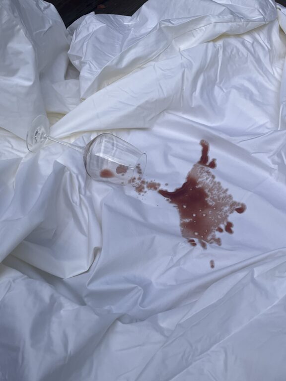 A clear wine glass tipped over on a white sheet, spilling red wine and leaving a prominent stain.