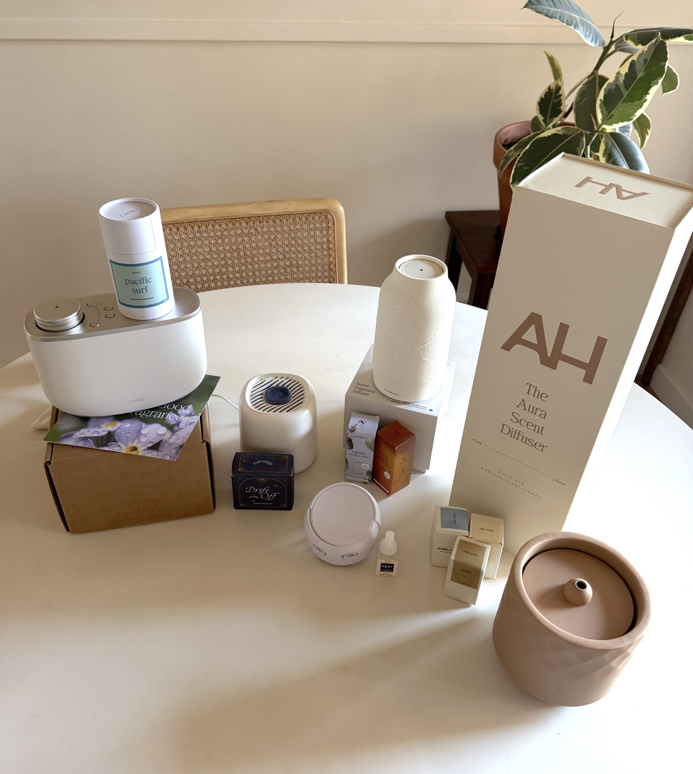 A variety of essential oil diffusers, scent cartridges, and fragrance boxes are arranged on a round white table with a chair and a plant in the background.