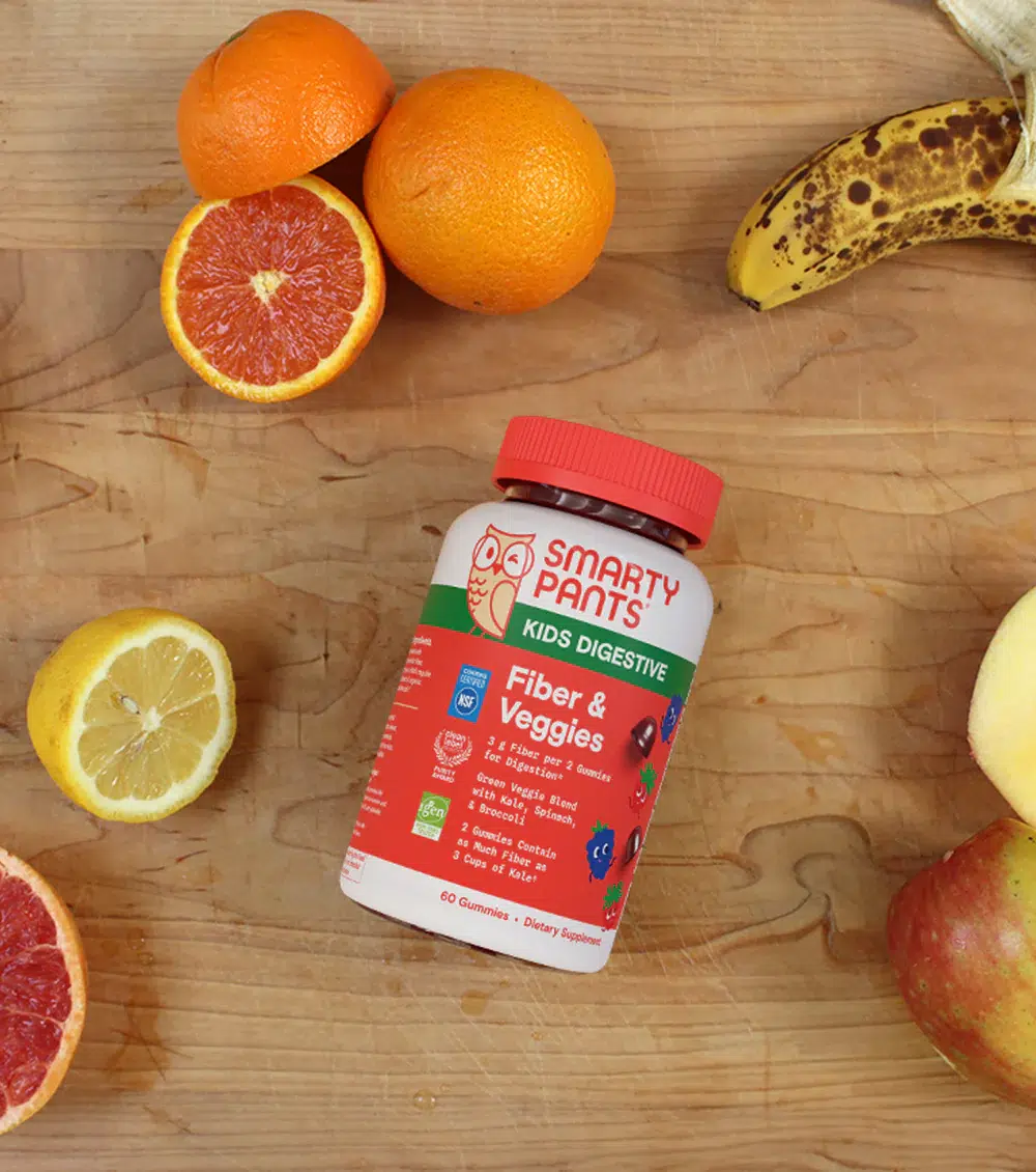A bottle of SmartyPants Kids Digestive Fiber & Veggies gummies on a wooden surface, surrounded by assorted fruits including orange, banana, lemon, grapefruit, and apple.