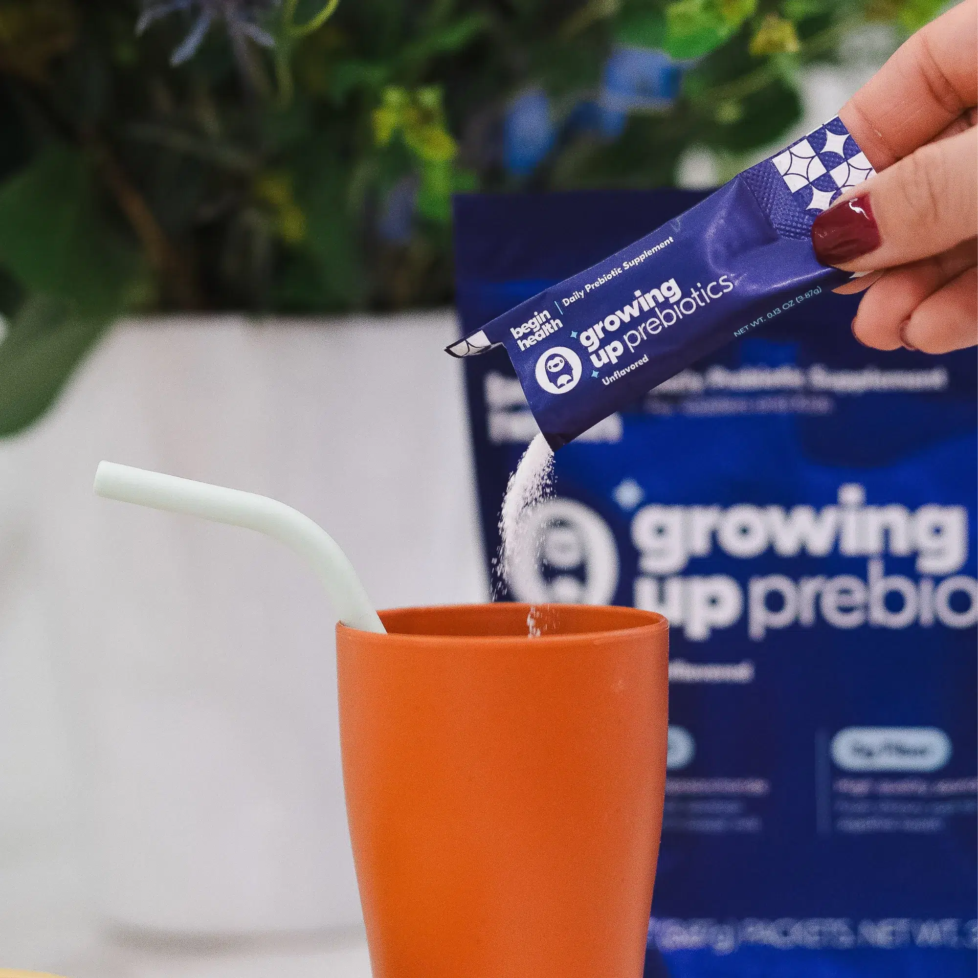 A hand pours a packet of "growing up prebiotics" powder into an orange cup with a white straw; a matching product package is in the background.