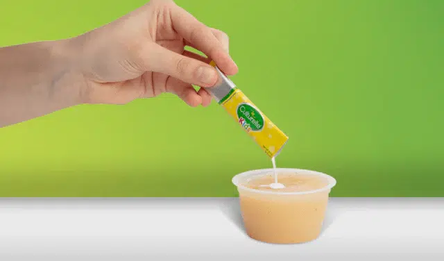 A hand pours a small tube of Amul condensed milk into a plastic container filled with a light brown liquid, with a green gradient background.